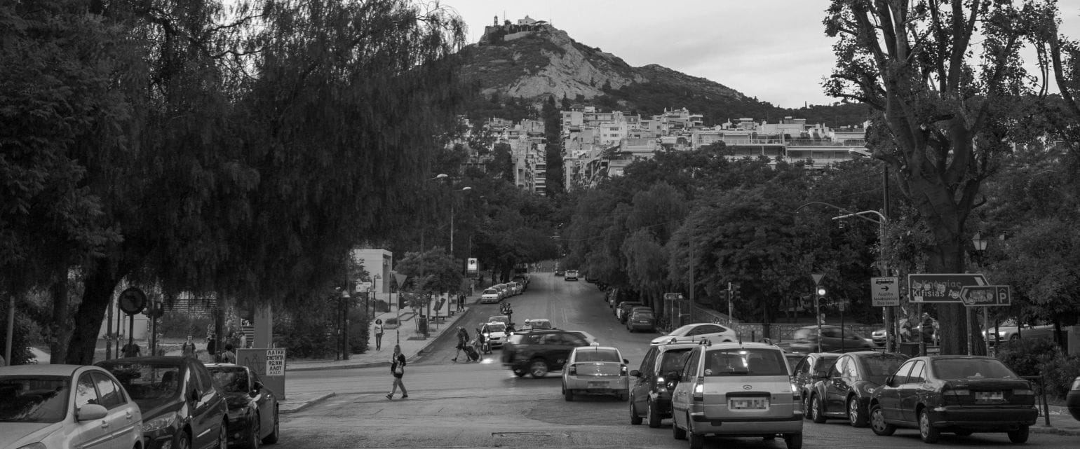 ασπρόμαυρη εικόνα Αθήνας, αυτοκίνητα στον δρόμο, στο φόντο δέντρα, σπίτια και ο Λυκαβηττός