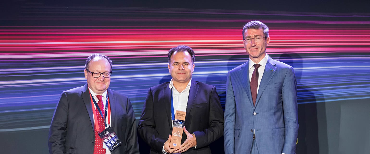Τρεις άνθρωποι. Στη μέση ο Αλέξης Πανταζής κρατάει το βραβείο. Στο φόντο neon κόκκινες, λευκές και μπλε γραμμές