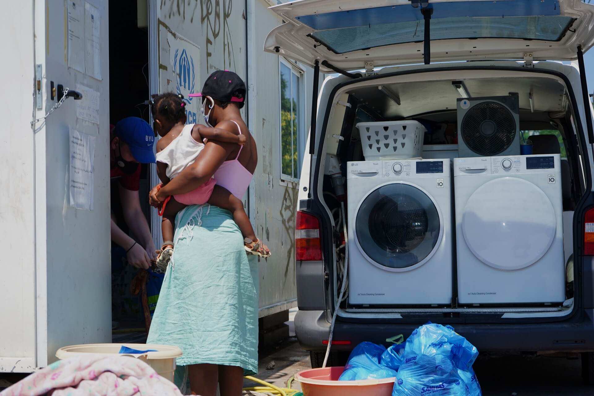 γυναίκα κρατά μωρό στην αγκαλιά της, δίπλα της φορτηγό με φορητά πλυντήρια ρούχων