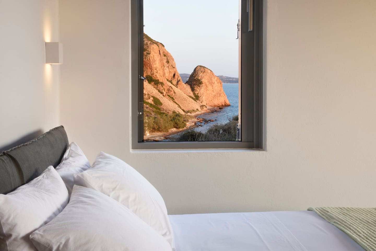 κρεβάτι με λευκά σεντόνια, θέα βράχος και θάλασσα μέσα από το παράθυρο