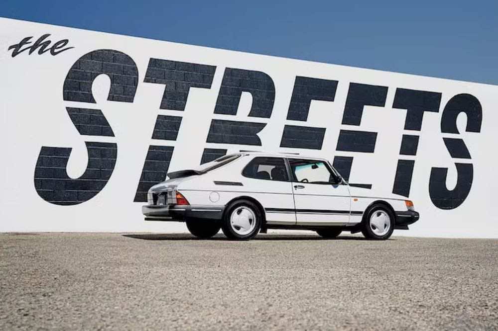 Saab άσπρο παρκαρισμένο μπροστά αό τοίχο με γραμμένες τις λέξεις the Streets