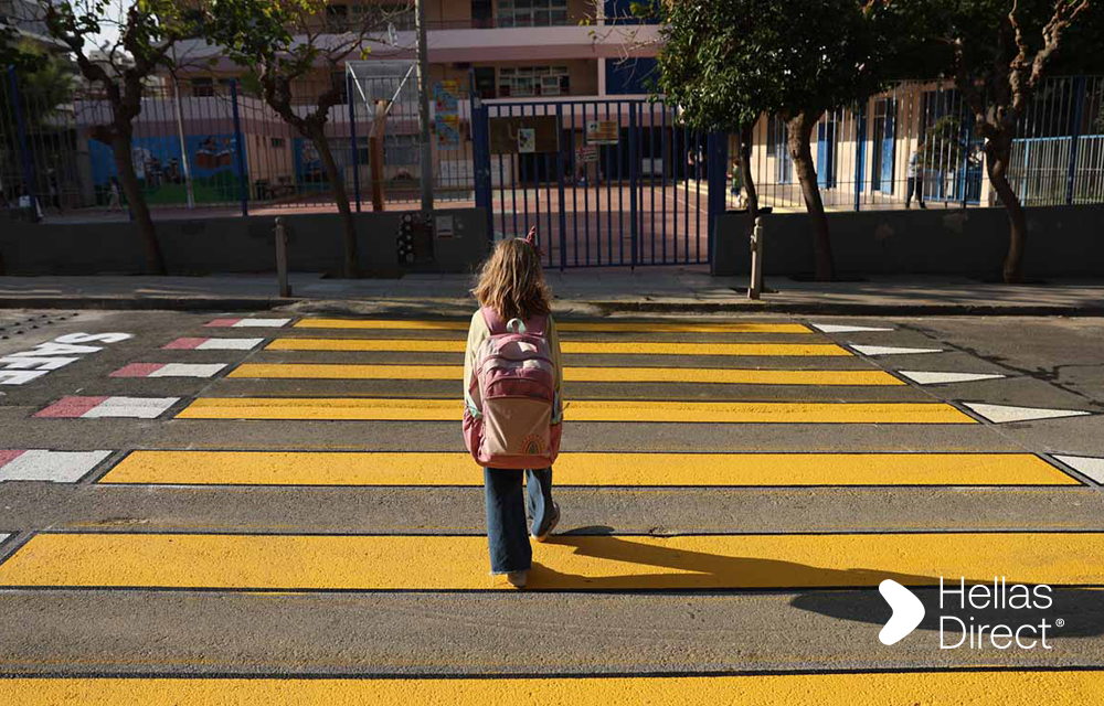 Δρόμος έξω από σχολείο, διάβαση με κίτρινα μολύβια αντί για λευκή διαγράμμιση, τη διασχίζει ένα μικρό κορίτσι που βλέπουμε πλάτη