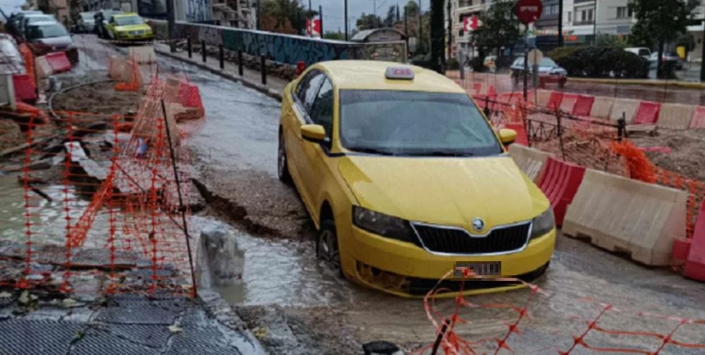 ταξί από μπροστά, έχει πέσει μέσα σε λακκούβα με νερά στην άσφαλτο, γύρω πορτοκαλί κορδέλες έργων