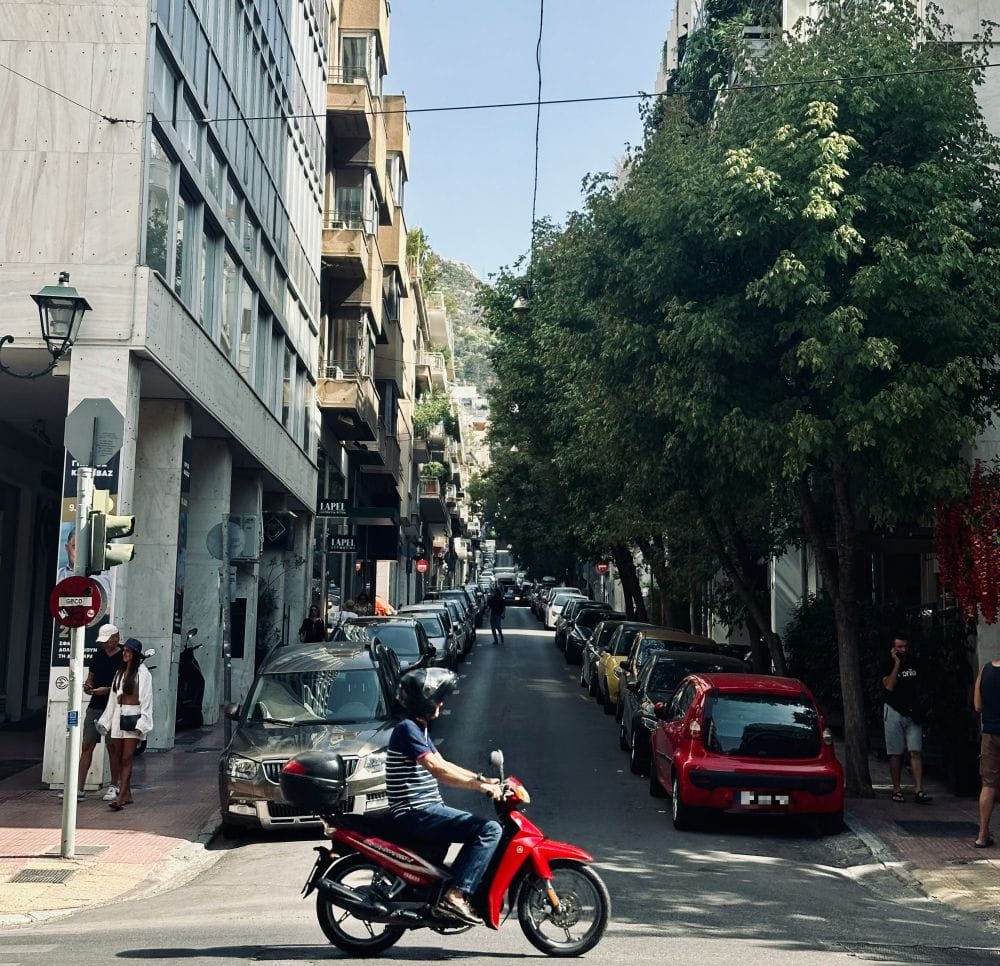 Εικόνα από το κέντρο της Αθήνας, δρόμος με παρκαρισμένα, κτίρια δεξιά αριστερά, μηχανάκι και πεζοί. Στο βάθος ο Λυκαβηττός