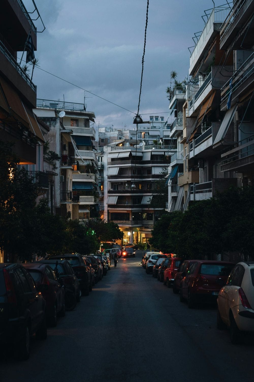 δρόμος της Αθήνας σούρουπο, παρκαρισμένα αυτοκίνητα δεξιά κι αριστερά, στο βάθος αυτοκίνητο με αναμμένα φώτα και γύρω γύρω πολυκατοικίες