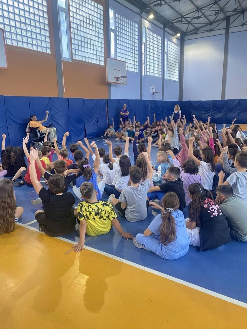 Ο Δημήτρης Αντωνίου σε κλειστό σχολικό γήπεδο, παιδιά καθισμένα κάτω τον ακούν και σηκώνουν τα χέρια τους