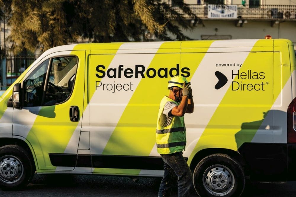 το βαν του Safe Roads project από το πλάι, μέλος του συνεργείου με κίτρινο γιλέκο και προστατευτικό κράνος περνά από μπροστά