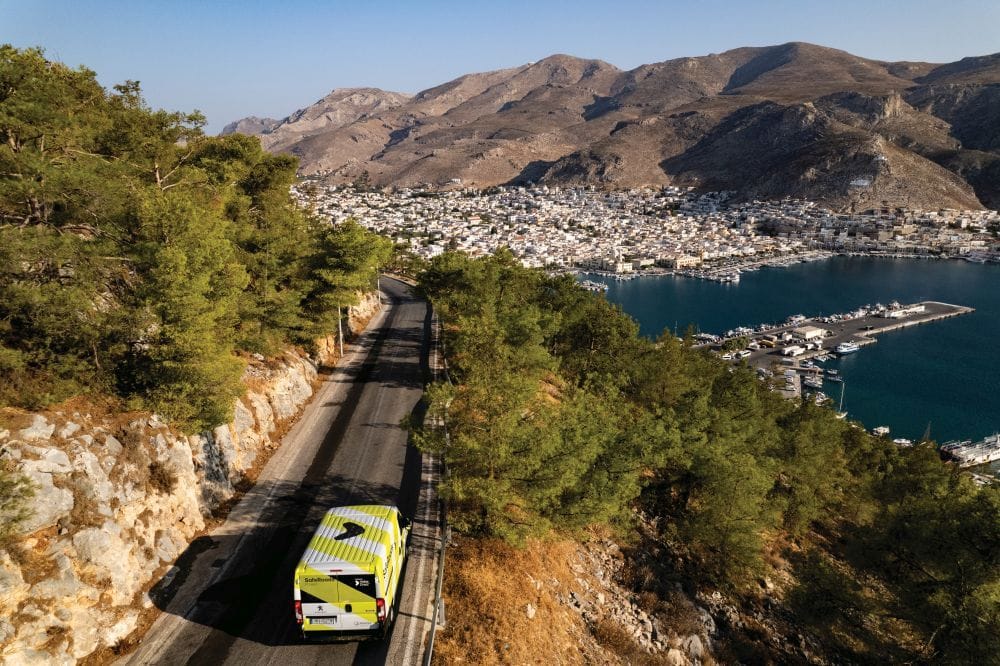 εικόνα νησιού από ψηλά στο φόντο λιμάνι με άσπρα σπίτια και βουνά, μπροστά το βαν του Safe Roads project στον δρόμο