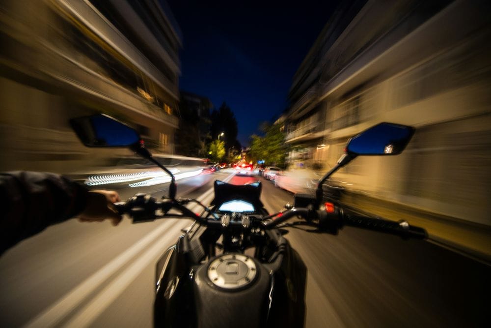 οπτική οδηγού μηχανής που κοιτάει μπροστά, φαίνεται το φέρινγκ, το τιμόνι και οι καθρέφτες. Μπροστά δρόμος με αυτοκίνητα και φώρα, δεξιά κι αριστερά σπίτια