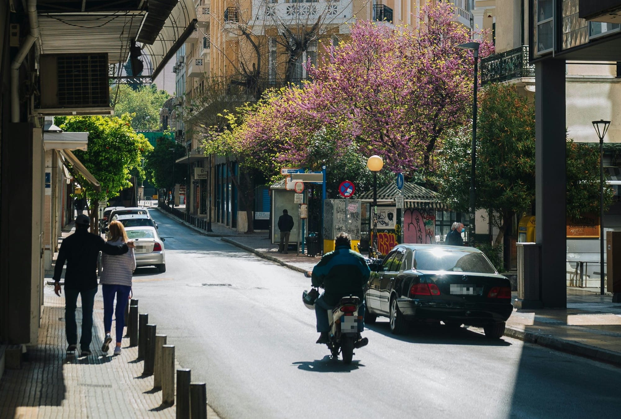 εικόνα πόλης, δρόμος με λίγα παρκαρισμένα αυτοκίνητα, στη μέση μηχανάκι από ίσω, ο οδηγός κρατάει το κράνος στο χέρι. Δεξιά κι αριστερά πεζοδρόια και κτίρια.
