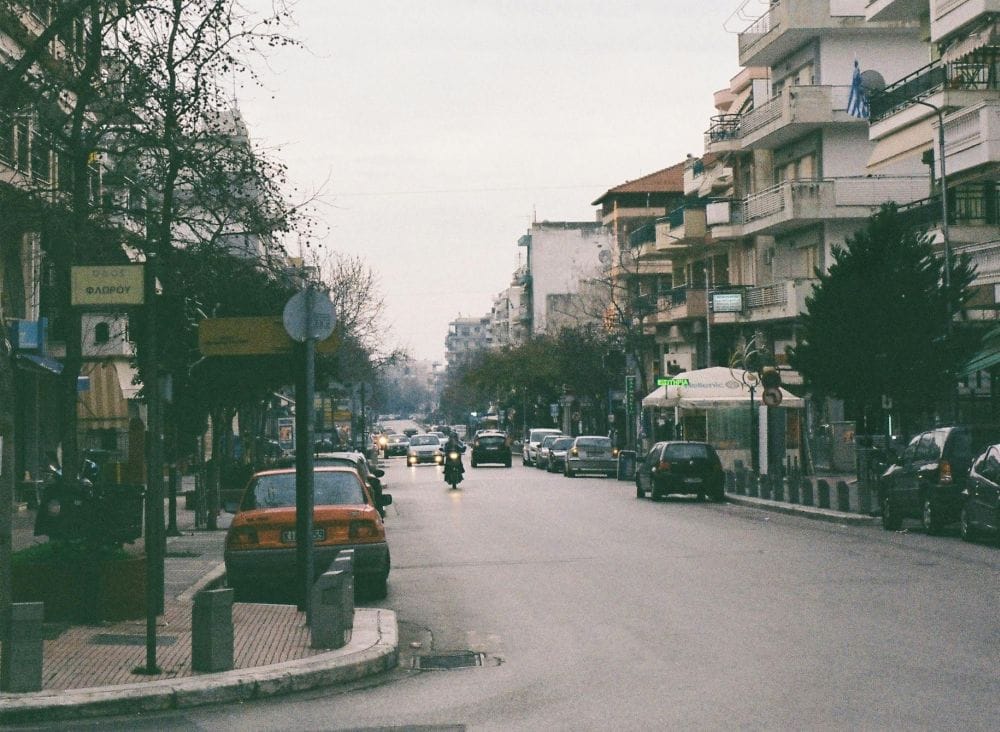 εικόνα δρόμου πόλης, άσφαλτος στη μέση, αυτοκίνητα παρκαρισμένα και εν κινήσει, περίπτερο και κτήρια δεξιά αριστερά
