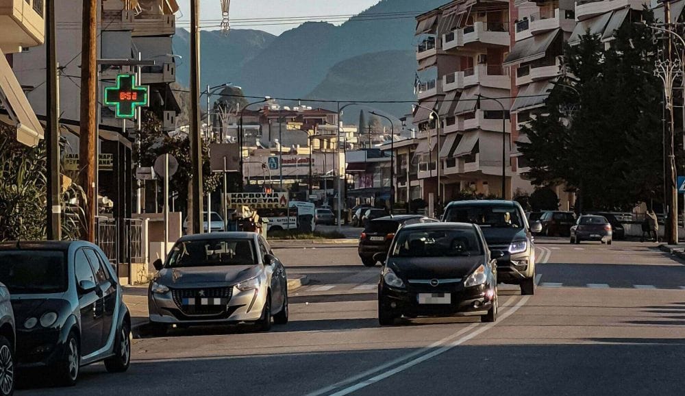 δρόμος πόλης μέρα, αυτοκίνητα εν κινήσει και παρκαρισμένα, δεξιά κι αριστερά πολυκατοικίες