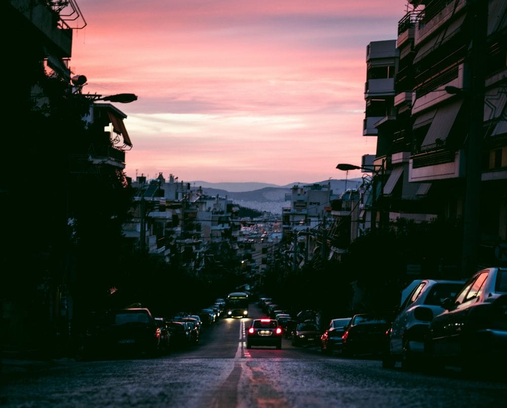 δρόμος πόλης με αυτοκίνητα παρκαρισμένα και εν κινήσει για το θέμα τι καλύπτει η ασφάλεια αυτοκινήτου για αλκοόλ και οδήγηση. Δεξιά κι αριστερά πολυκατοικίες, στο φόντο ουρανός στο ηλιοβασίλεμα