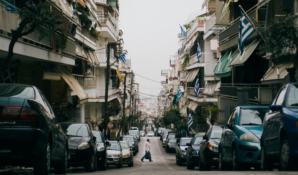 εικόνα δρόμου για το θέμα: τεκμήρια αυτοκινήτων. Δεξιά κι αριστερά πολυκατοικίες και παρκαρισμένα αυτοκίνητα, στη μέση πεζός διασχίζει τον δρόμο