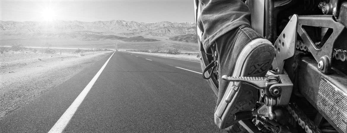 ασπρόμαυρη εικόνα, πόδι οδηγού πάνω σε πενταλ μηχανής, μπροστά ο ανοιχτός δρόμος και στο φόντο βουνά