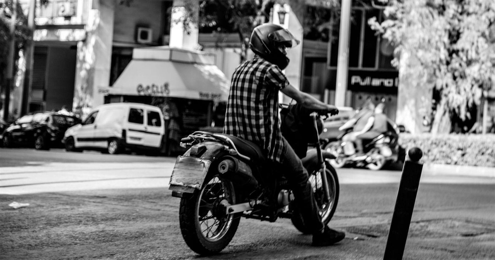 ασπρόμαυρη εικόνα, οδηγός μηχανής σταματημένος σε δρόμο της Αθήνας, γύρω κτίρια και δέντρα