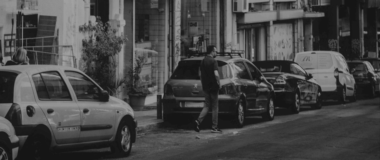 Ασπρόμαυρη εικόνα πόλης, δρόμος με παρκαρισμένα αυτοκίνητα στη σειρά, άνθρωπος περπατά δίπλα τους