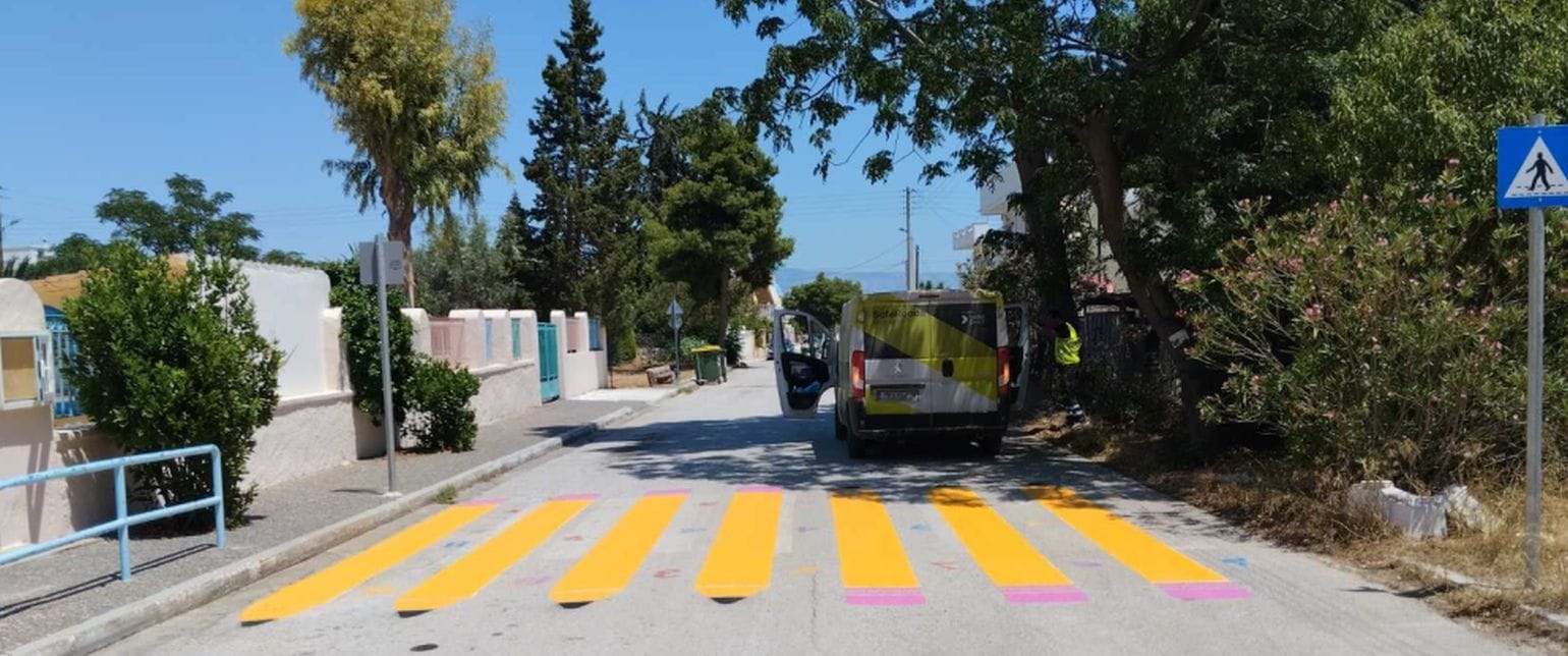 δρόμος με χαμηλά κτίρια αριστερά και δέντρα δεξιά, στη μέση διάβαση με κίτρινα μολύβια και το βαν του Safe Roads project από πίσω