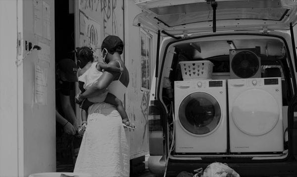 ασπρόμαυρη εικόνα, γυναίκα κρατά μωρό στην αγκαλιά της, δίπλα της φορτηγό με φορητά πλυντήρια ρούχων