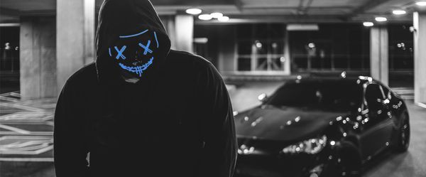Ασπρόμαυρη εικόνα με μπλε στοιχεια, μπροστα άνδρας με κουκούλα και μάσκα, πίσω του παρκαρισμένο μαύρο αυτοκίνη
