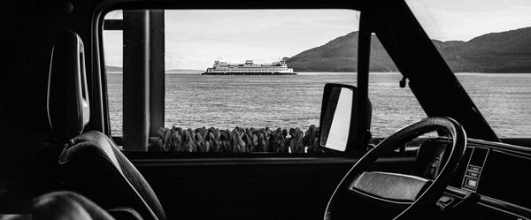 ασπρόμαυρη εικόνα μέσα από αυτοκίνητο, φαίνεται από το πλάι κάθισμα οδηγού και τιμόνι και στο βάθος θάλασσα κι ένα πλοίο